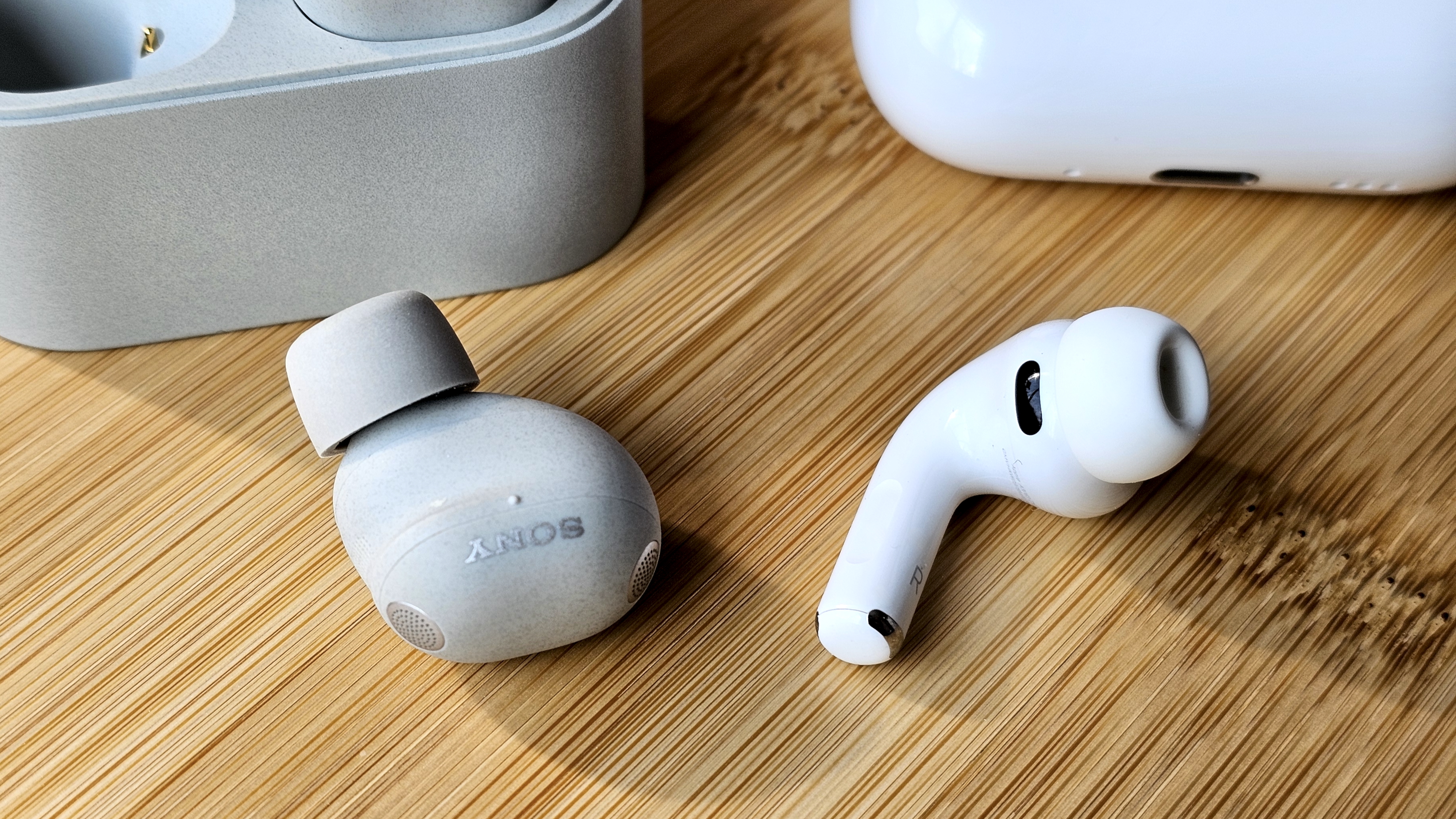 Sony WF-1000XM6 earbuds next to the Apple AirPods Pro 3, on a wooden table, in a versus-style format
