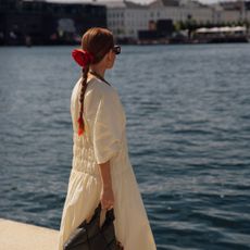 a woman stands looking at the Copenhagen canal wearing a red rosette hair clip and an ivory dress with a black leather bag