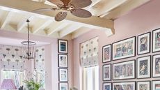 Pale pink dining room with white beams, a gallery wall of prints, palm tree printed fabric blinds, and a rattan ceiling fan