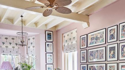 Pale pink dining room with white beams, a gallery wall of prints, palm tree printed fabric blinds, and a rattan ceiling fan