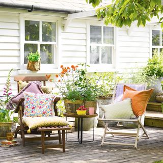 Two deckchairs with colourful cushions on decking by the white wooden clad house with pots of plants