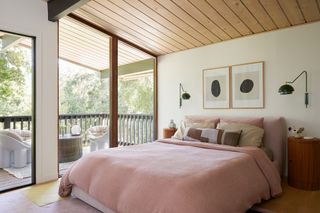 japandi style bedroom with timber panelled slanted ceiling, large windows, artwork hanging over the bed, two wall sconces, timber side table, and a pastel pick bedspread