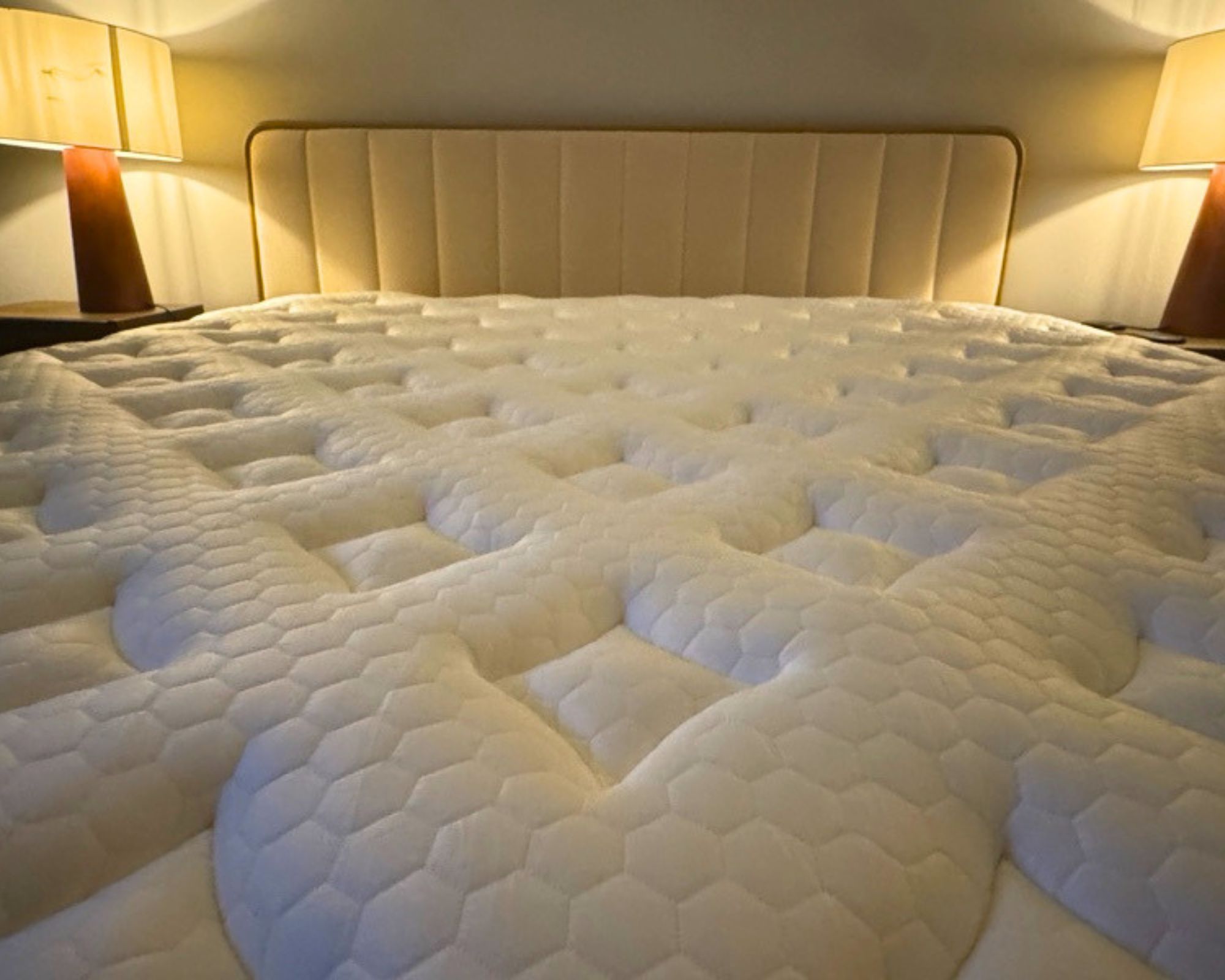 A close-up of the Nolah Evolution mattress shown on a bed with a fluted white and wood headboard, with two matching red lamps to either side against a white wall.