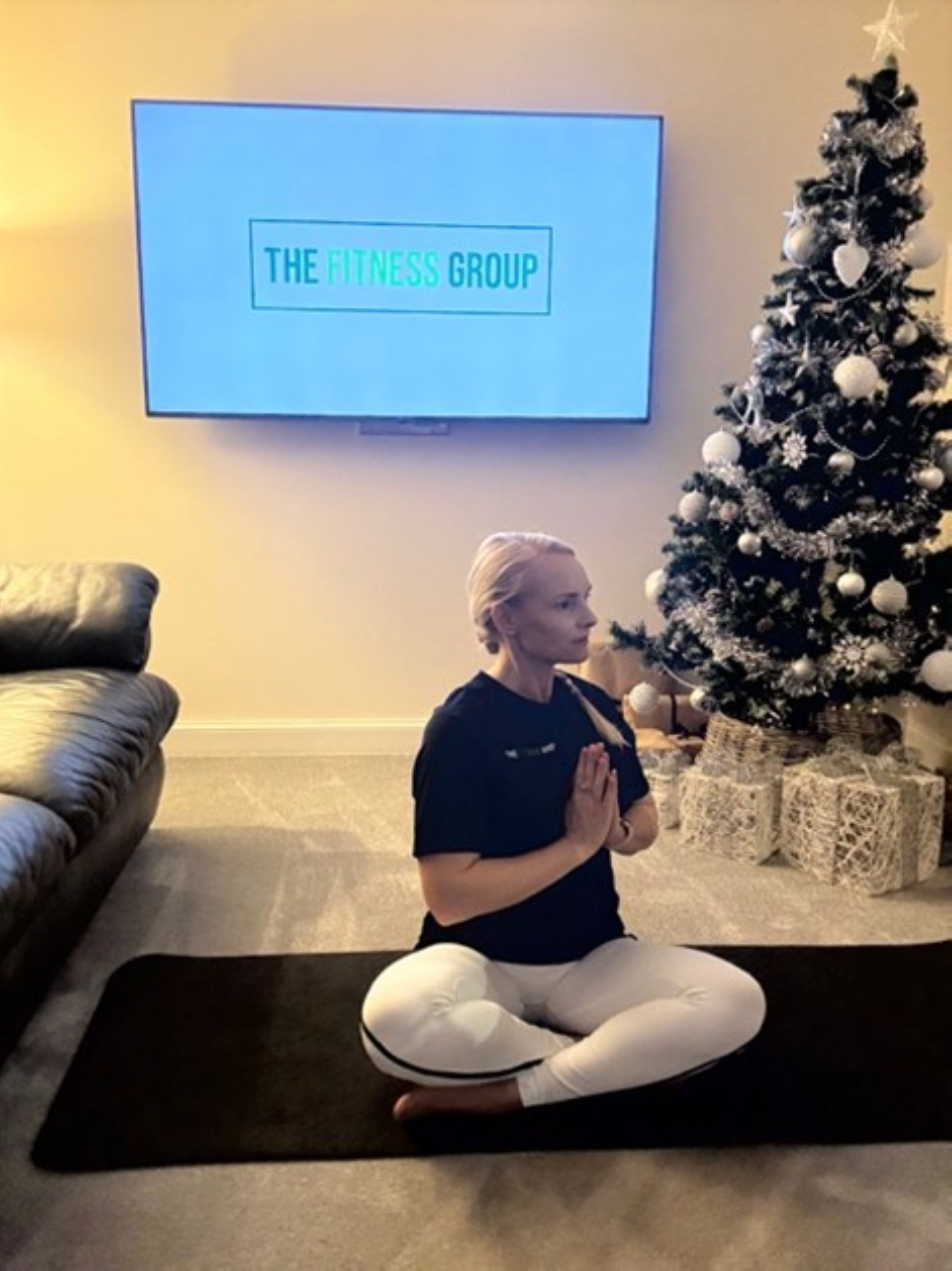 Woman demonstrates a Pilates exercise between a sofa and a Christmas tree. A mounted TV in the background reads "The Fitness Group"
