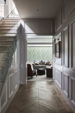 Hallway with wall paneling, chevron parquet flooring, staircase with clear baluster and view to living room