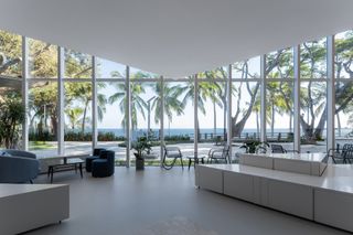 White interior room with table, chairs and lounge seating, white glossed triangular units, white floor, glass framed wall, view of surrounding landscape, palm trees, trees, sea in the distance and blue sky