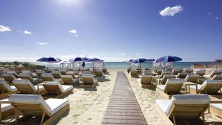 Beach image, white sand, wooden pathway, wooden sunloungers with white cusgions, blue and green parasols, blue sea, blue sky with small white clouds