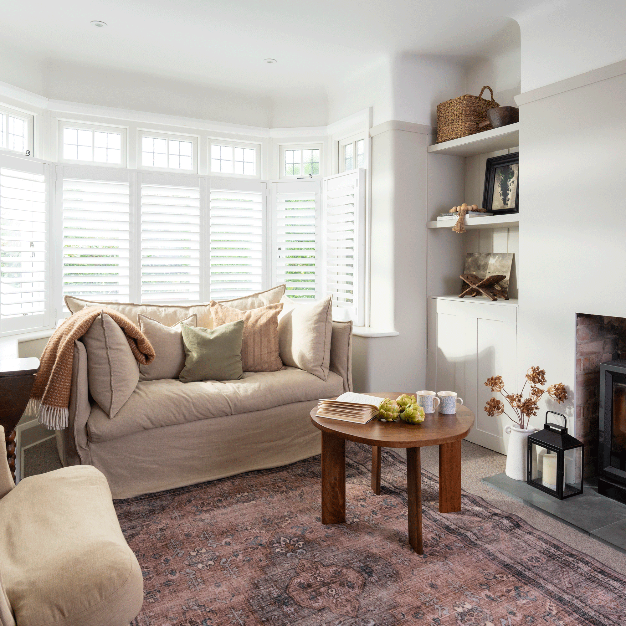 a neutral living room with a faded pink rug and beige upholstered seating with shutters on the window