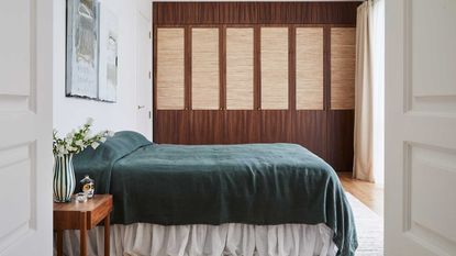 A bedroom with a minimalist, emerald green coverlet and a white ruffled bed skirt. The walls are white except for the back wall, which is a mix of dark and light wood paneling. There is a wooden bedside table with a green and white striped flower vase on it.