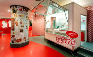 Currywurst museum, red and grey design floor and walls, white ceiling, currywurst stall, serving hatch open and lit up, column decorated with relevant food posters and articles