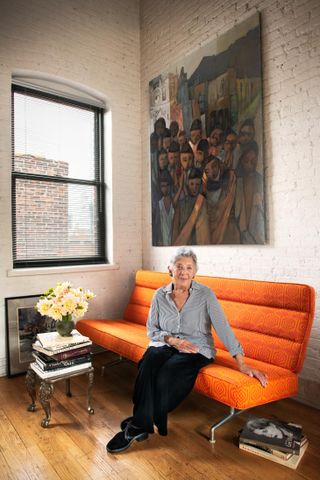A portrait of Lee Gross sitting on an orange sofa in her Manhattan apartment