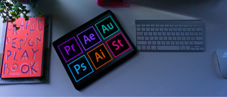 A tablet showing Adobe app icons next to a computer and keyboard and graphic design desk 