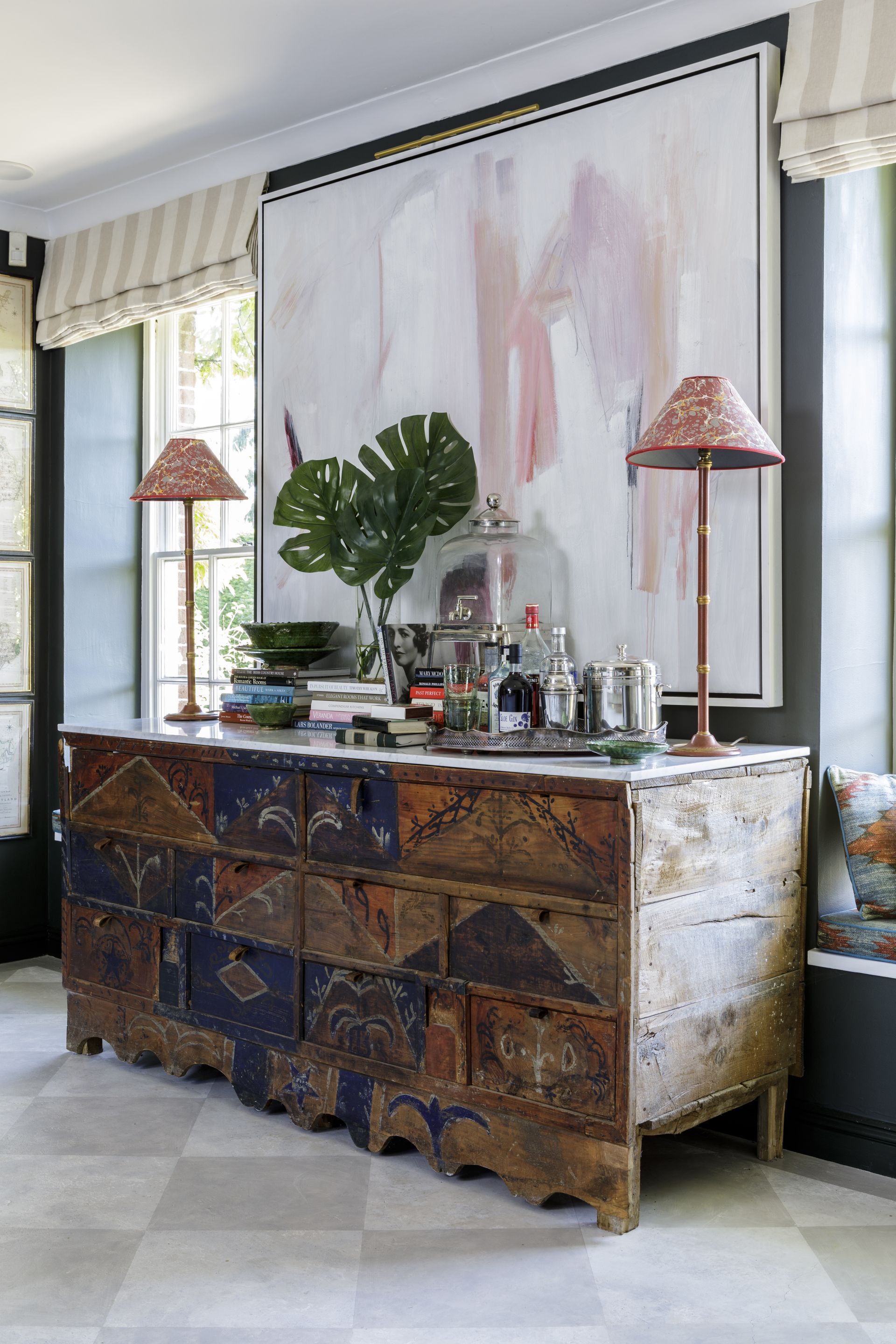 Wooden chest of draws with blue and red hand painting design, grey walls with window seats and large white painting