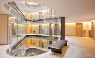 Interior view of floor five at The One, Barcelona, Spain featuring a dark coloured bench style seat, light coloured walls and floor, doors with small digital screens beside them and a large clear curved barrier that continues through other floors