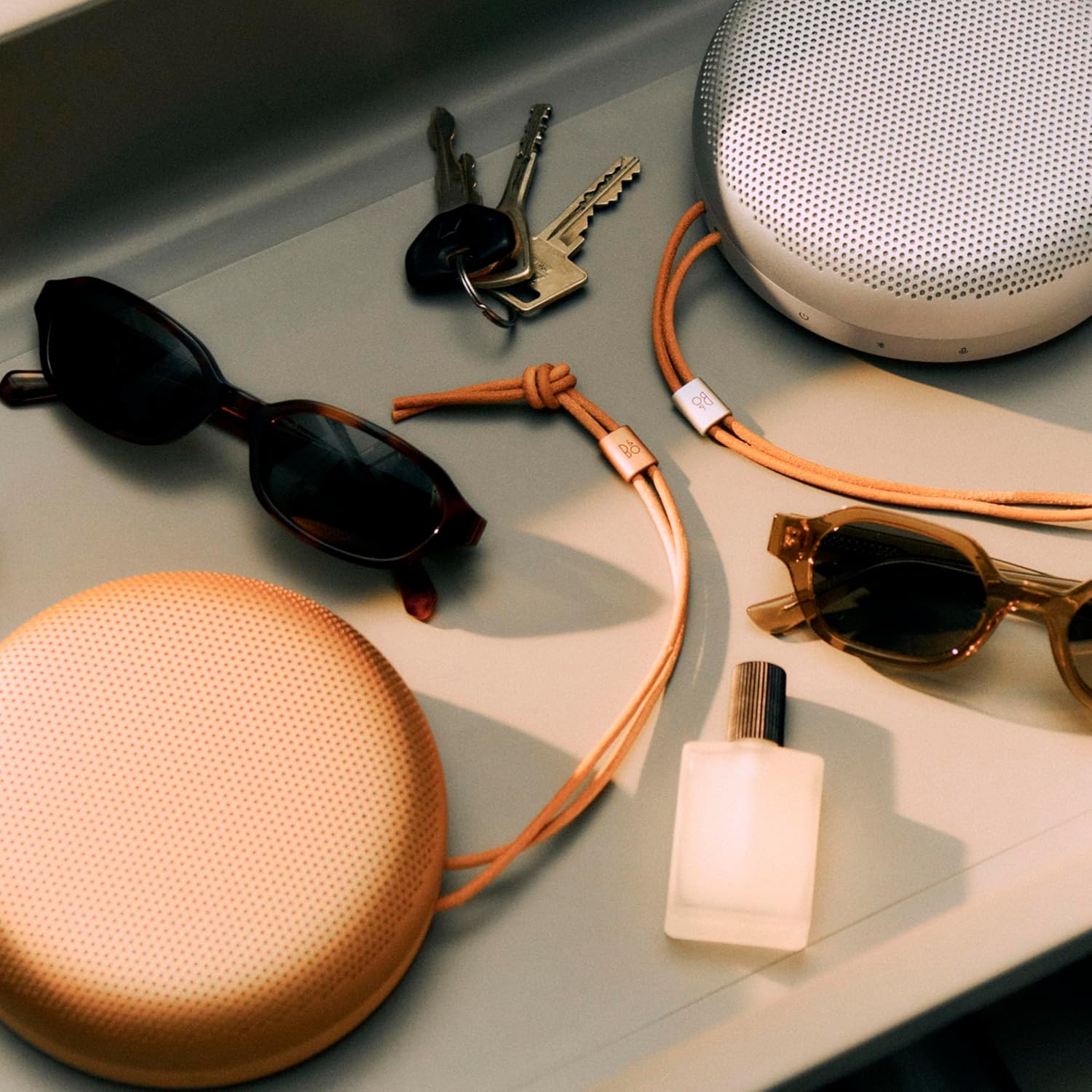Image of a tray that has two stylish, metal, portable speakers on it as well as two pairs of sunglasses, keys, and a perfume bottle.