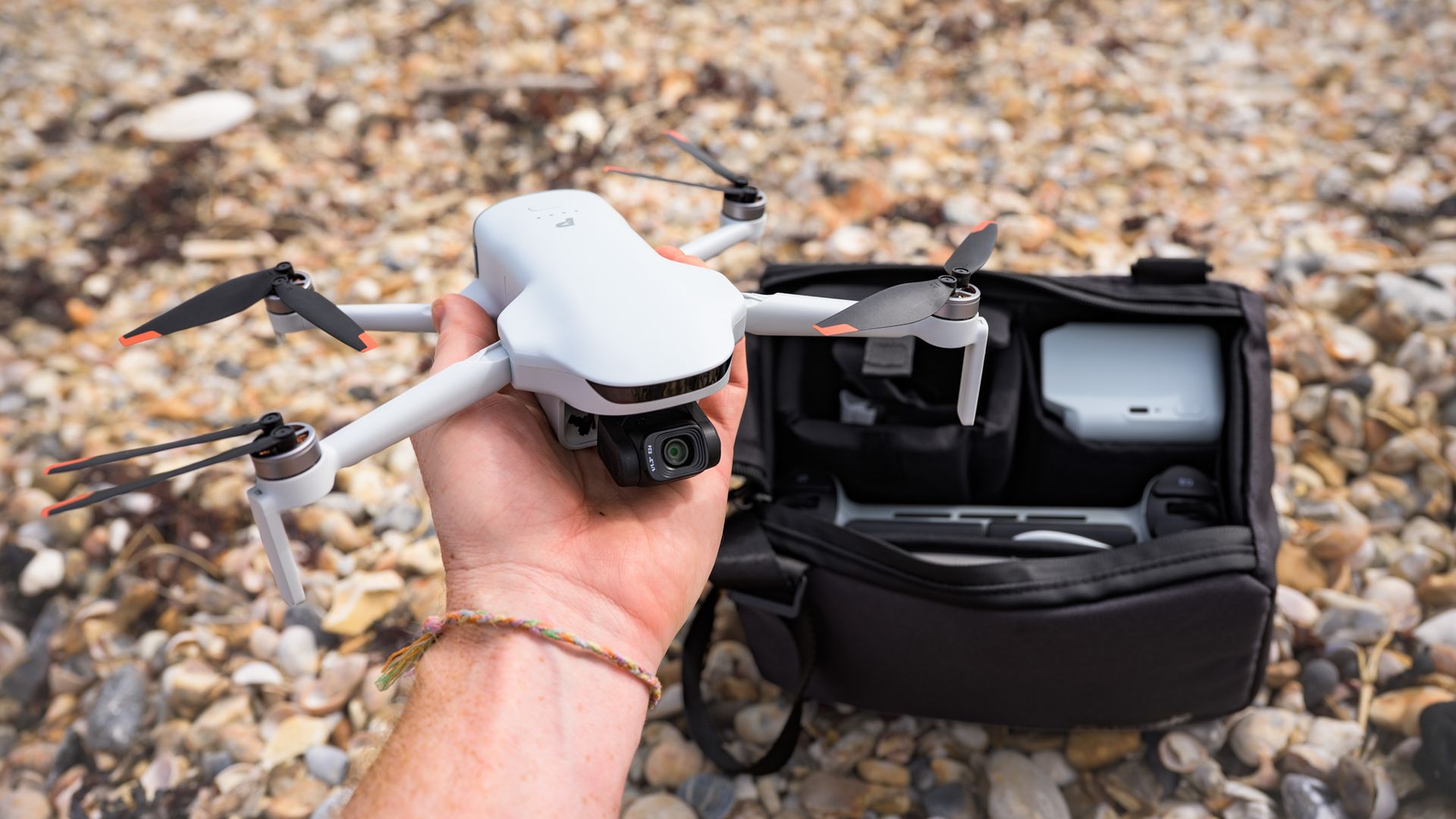 Potensic Atom 3 beginner drone in user's hands and the Fly More Combo bag and its content behind it, on a pebble beach