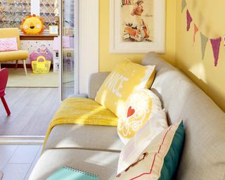 A living room with yellow wall paint decor, grey sofa, bunting and kids playroom in background