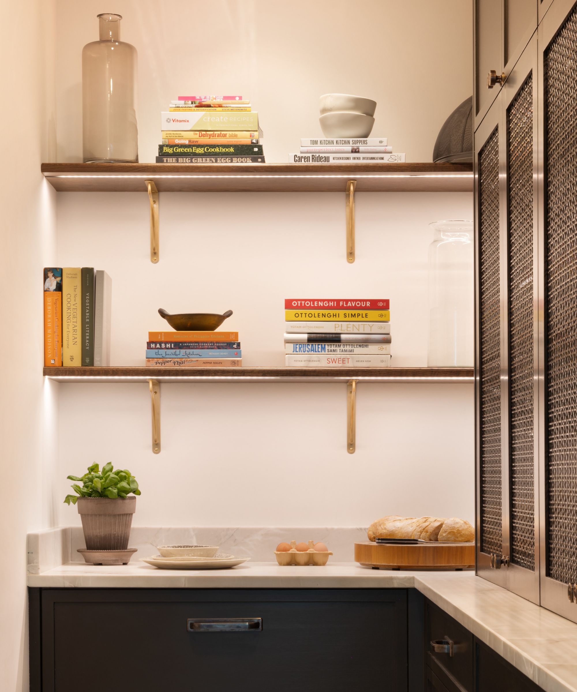 A compact kitchen nook with two wooden shelves mounted on white walls, each supported by brass brackets. The shelves are stacked with colourful cookbooks, bowls, and jars. Below, a marble countertop holds a potted basil plant, a plate, a carton of eggs, and a loaf of bread on a wooden board.