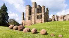 Eight stone catapult shots sit in the grass in front of a medieval castle. 