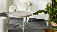 A beige mesh outdoor coffee table with a bottle and glass of water on top in front of two white outdoor chairs on a dark tiled floor with foliage on one side and an outdoor closet on the other