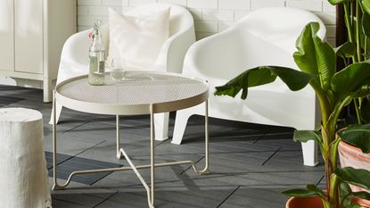 A beige mesh outdoor coffee table with a bottle and glass of water on top in front of two white outdoor chairs on a dark tiled floor with foliage on one side and an outdoor closet on the other