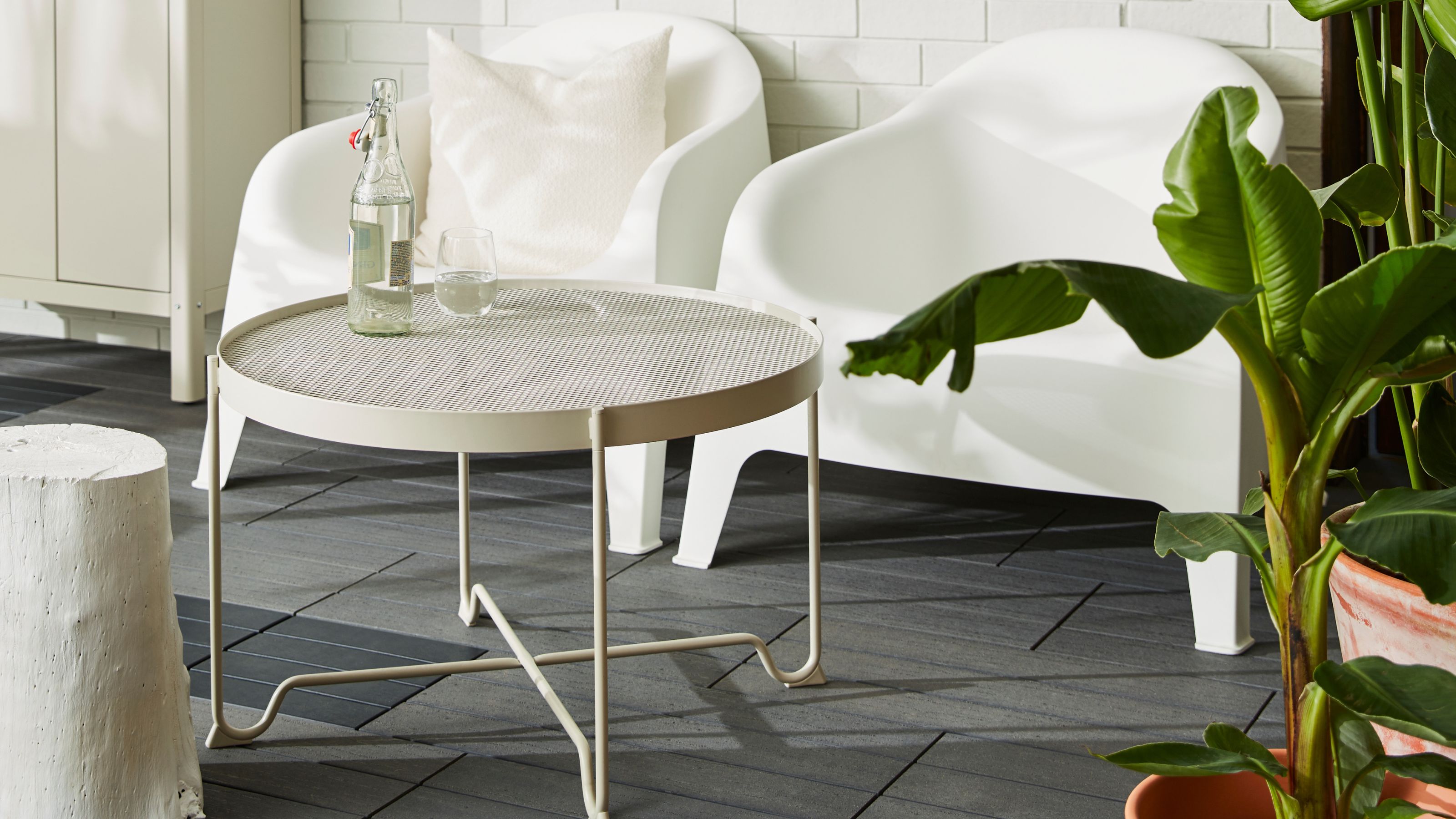 A beige mesh outdoor coffee table with a bottle and glass of water on top in front of two white outdoor chairs on a dark tiled floor with foliage on one side and an outdoor closet on the other