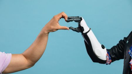 A person's left hand comes from the left of the image to meet a black and white robotic hand from the right of the image to make a heart with their hands in the center, all in front of a blue background.