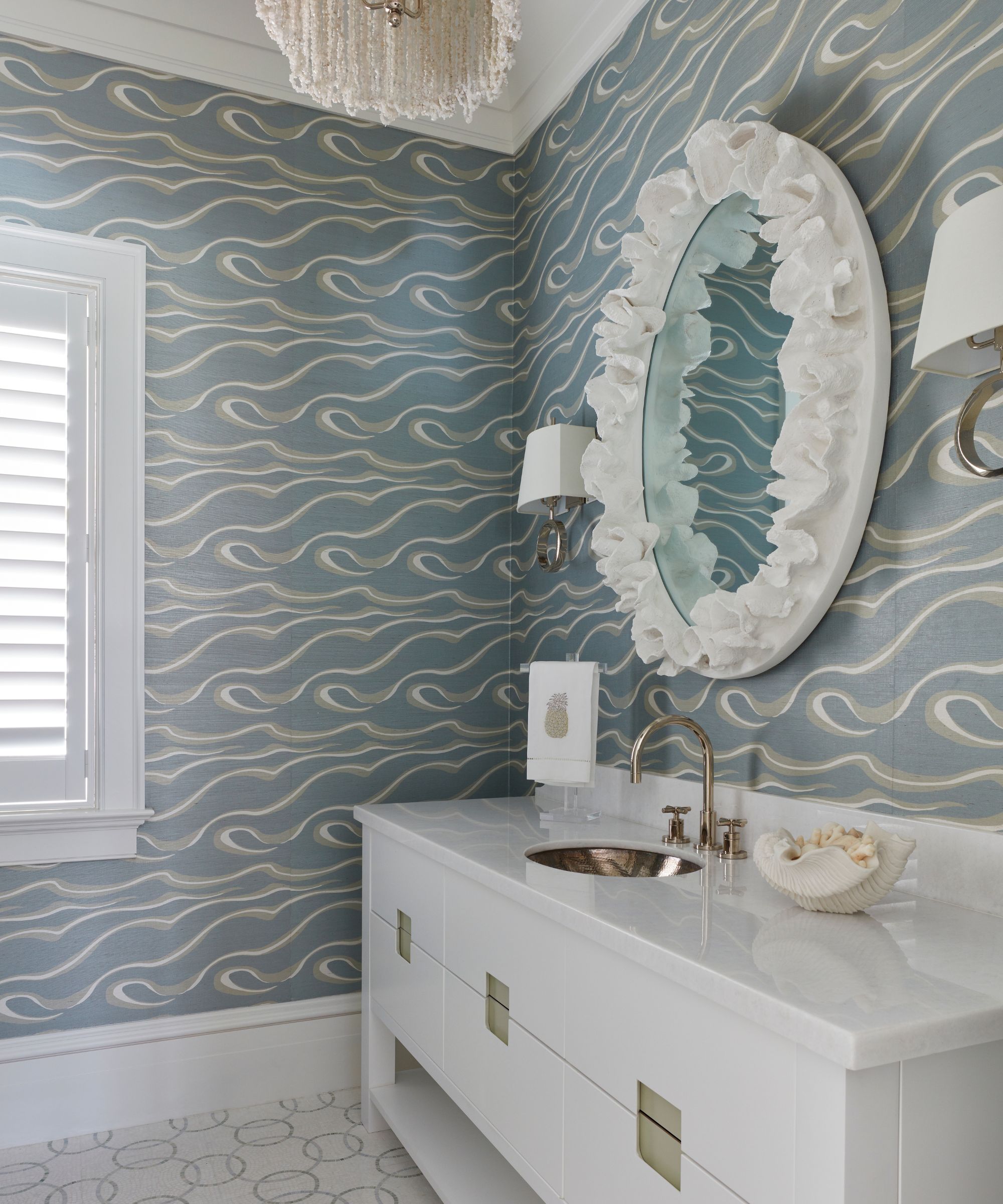 A coastal bathroom with blue wallpaper, a white vanity, and a decorative round white wall mirror