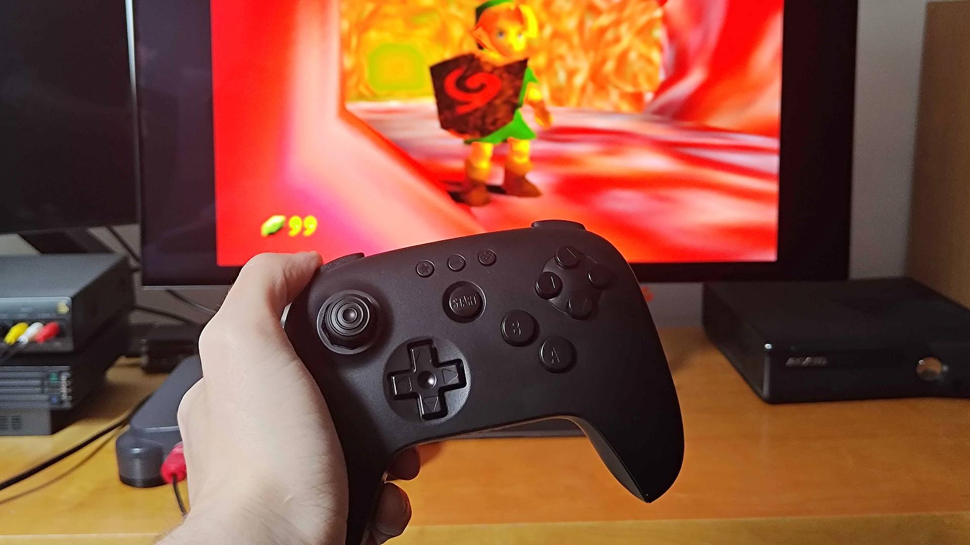 Hand holding 8Btido N64 controller with Zelda: Ocarina of Time gameplay on screen in backdrop.
