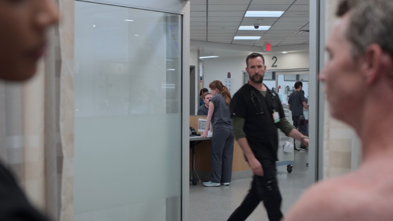 Dr. Robby walking past an open door while Dr. Mohan is helping Dr. Abbot on The Pitt.