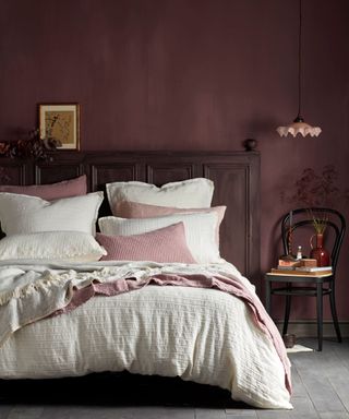 Bedroom with dark pink painted walls, looking at double bed with white bedding, pink blankets and scatter cushions, low hanging pendant above black wooden chair decorated with books, flowers and ornaments, dark wooden headboard, gray-brown flooring, framed picture and foliage resting on headboard.