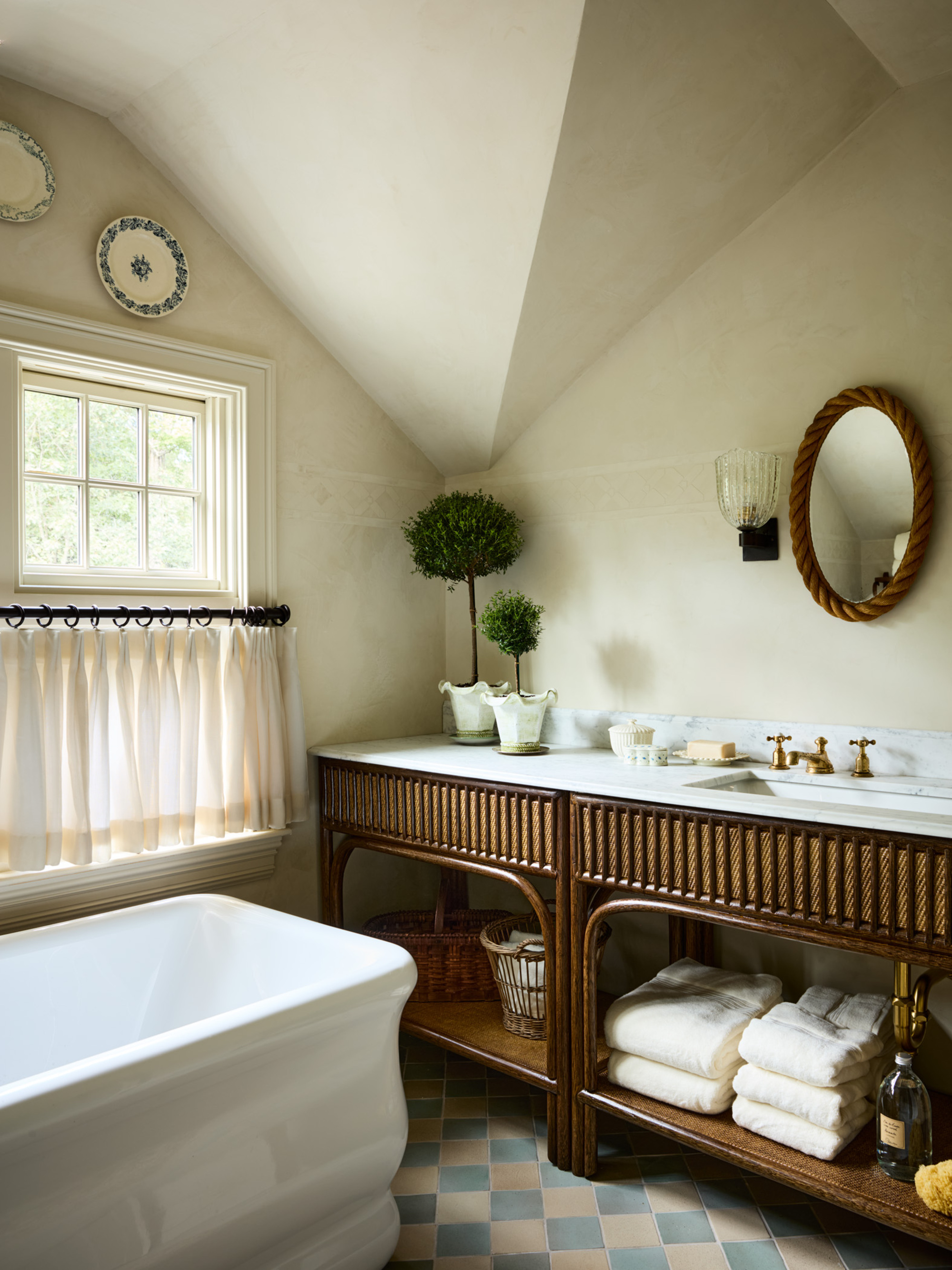 Image of a bathroom with off-white stone walls and a large sink unit that has a marble top and a wooden bottom with open shelves. There is a small rope mirror, a window with cafe curtains, and a tub.