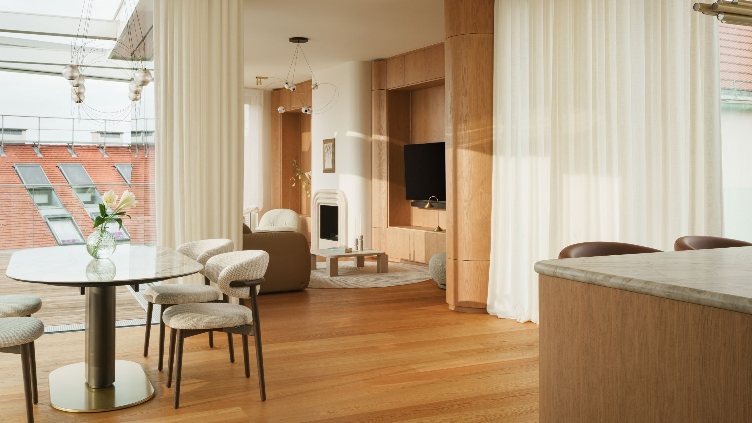 Image of an open plan home with a kitchen, dining area, and living room all in view. There are wooden floors, wooden walls, large white curtains, and lots of curved details. 