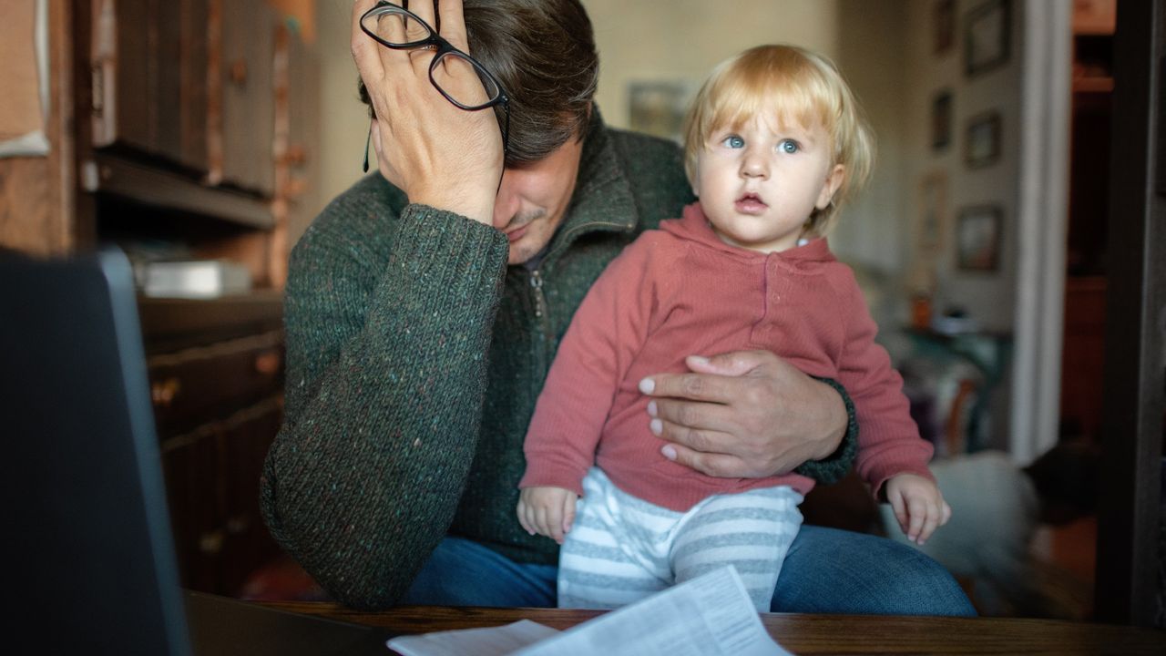 Worried man holding a young child at a desk looking over bills, worried about financial cost of childcare 