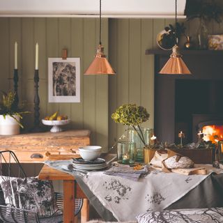 A wood-panelled green-painted dining room with two brass pendant lights hanging above the dining table and a wood-burning stove in the background