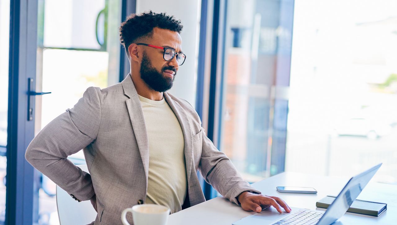 Man working at office desk sits uncomfortably, arching his back with one hand holding his lower back, a pained expression on his face. His other hand on the trackpad of a laptop on the desk. He wears a light-colored T-shirt and a taupe sports jacket.