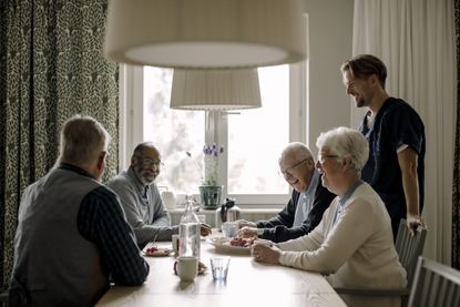 Cheerful elderly woman and men talking with male caregiver having food at table