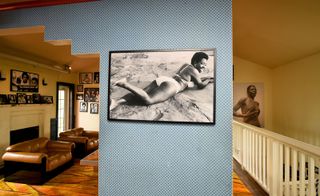A large black & white photo of a black female model is hung on the wall a light blue and white dotted wall. We see other photographs of the same nature through an opening, with light brown leather furniture.