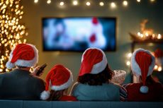 A family of four seen from behind watches Christmas movies on the couch while wearing Santa hats.