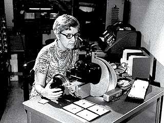 A black and white image of a woman behind a microscope.