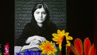 A lone silhouetted figure stands before a giant projected black-and-white portrait of Malala Yousafzai, surrounded by colourful animated flowers and foliage at the base of the image.