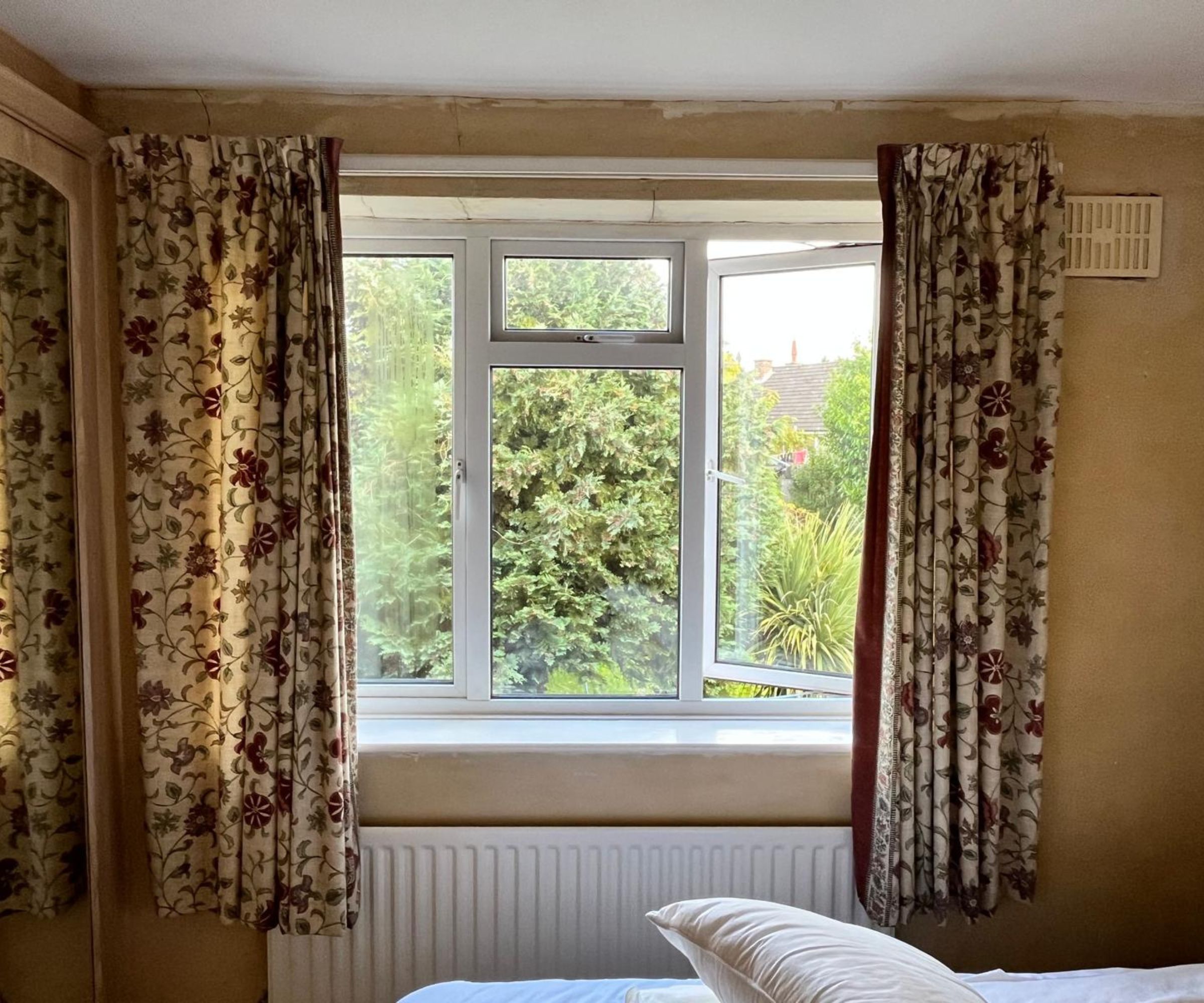 A dated bedroom with bare plaster walls, floral red and cream curtains framing a white uPVC window overlooking a green garden.