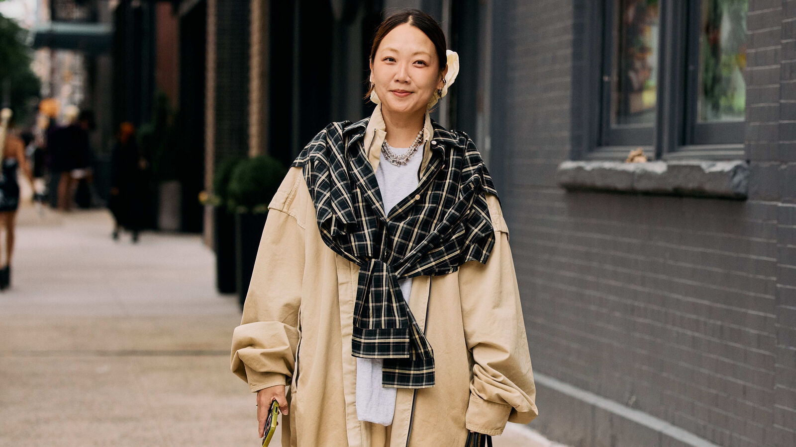 Woman wears oversized bomber, khaki trouser shorts, button down, plaid scarf, black handbag, and sandals. 