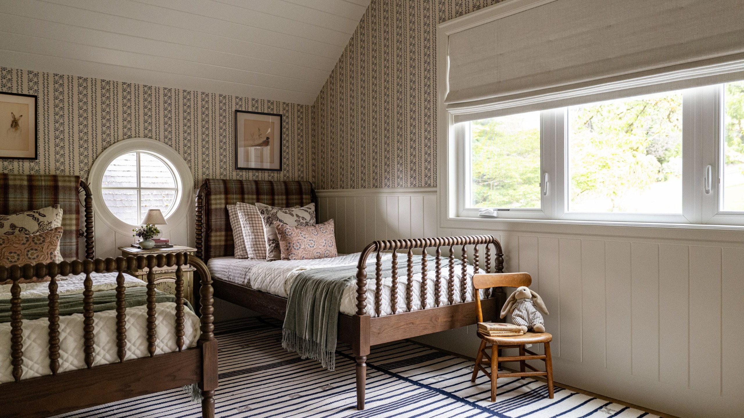 A kids' bedroom with twin spindle beds with plaid fabric headboards. A striped rug, warm white wall paneling, vintage-inspired wallpaper on the upper walls, and a small round window between the beds.
