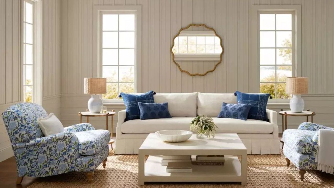 A white sofa with blue throw pillows and two blue floral armchairs clustered around a white coffee table; in the background, white panelled walls, a circular mirror, and windows. 