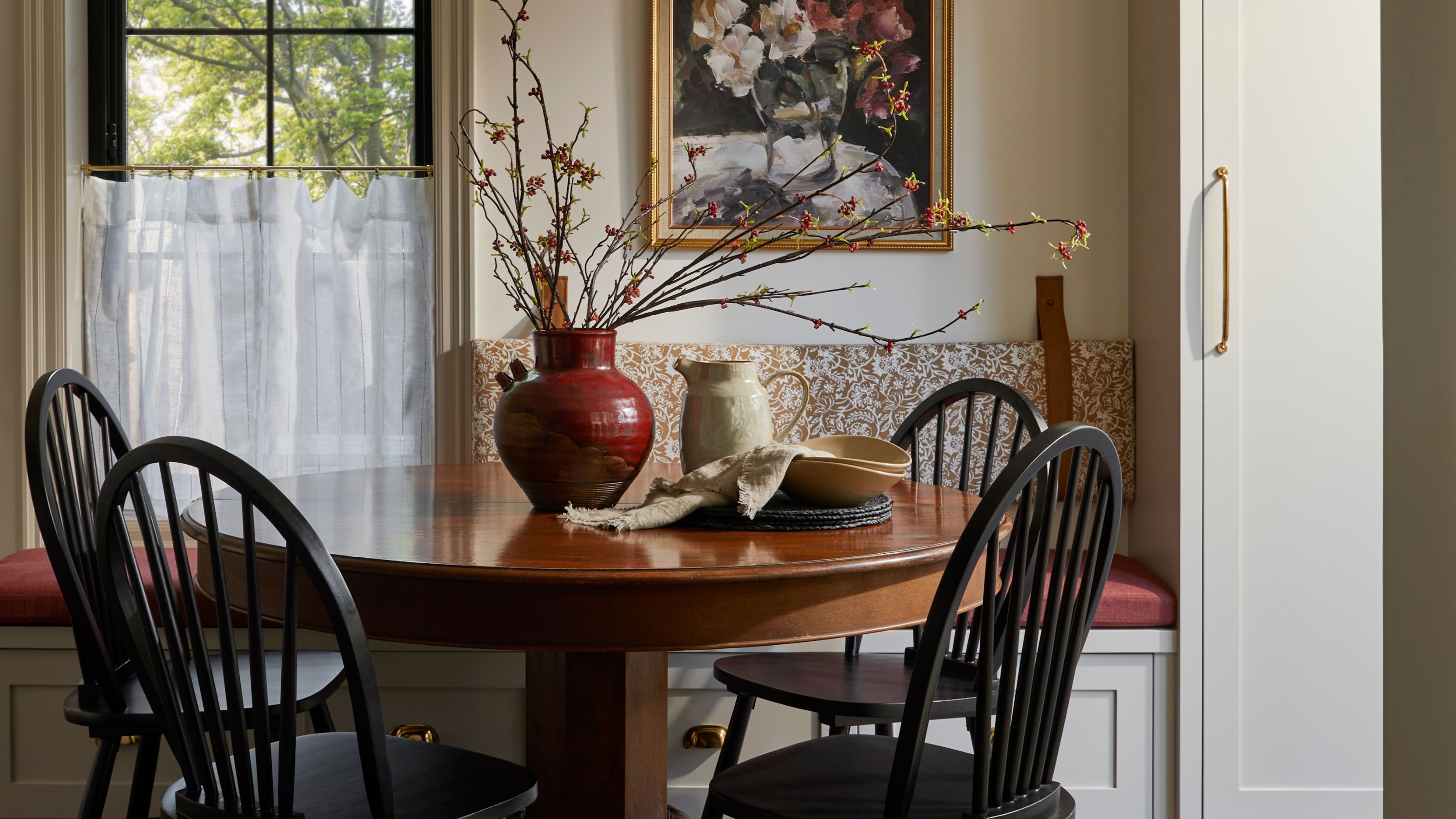 a cozy pattern layered dining nook with a banquette seat, small window and cafe curtain, wooden round table and black chairs