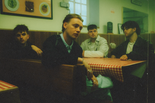 Four young men pose for a photo sat at a table in a dinner. This is Only The Poets, a British pop band.