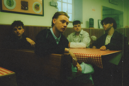 Four young men pose for a photo sat at a table in a dinner. This is Only The Poets, a British pop band. 