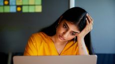A South Asian female employee wearing a bright orange shirt looks at her laptop with a stressed expression, with her head in her left hand. Behind her, a dark grey office wall with brightly colored sticky notes can be seen.