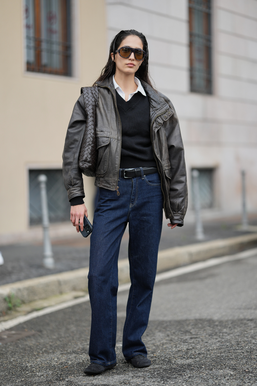 A guest wears long dark brown hair worn slicked back with a center part, oversized tortoiseshell gradient sunglasses, woven dark brown leather shoulder bag, white cotton collared shirt under a black knit v-neck sweater, dark brown leather zip-front jacket with flap pockets, black leather belt with silver buckle, dark indigo denim jeans in a straight-leg cut, black leather flat shoes, outside Tod&#039;s, during Milan Fashion Wee